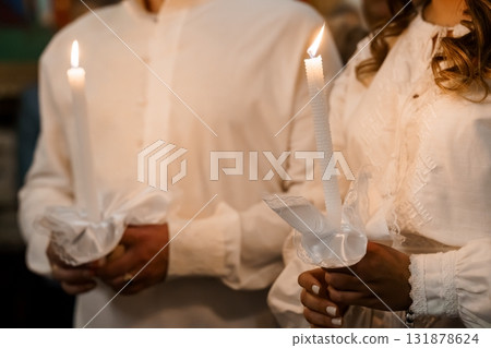 Orthodox Christian Wedding Ceremony: Couple Holding Lit Candles in White Robes, Church Interior, Warm Lighting, Religious Tradition. Orthodox Christian Wedding Ceremony: Couple Holding Lit Candles in White Robes, Church Interior, Warm Lighting, Religious Tradition. 131878624