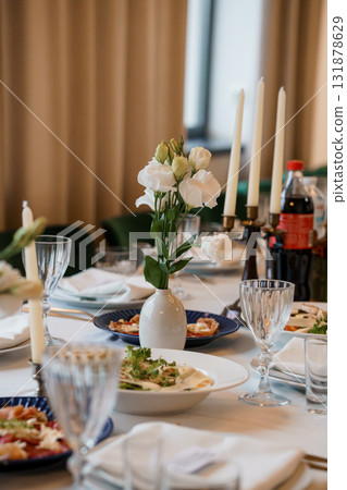 Elegant Dinner Table Setting with White Flowers in Vase, Candles, and Plates on Beige Linen. Stylish Food Presentation, Fine Dining Interior. Elegant Dinner Table Setting with White Flowers in Vase, Candles, and Plates on Beige Linen. Stylish Food Presentation, Fine Dining Interior. 131878629