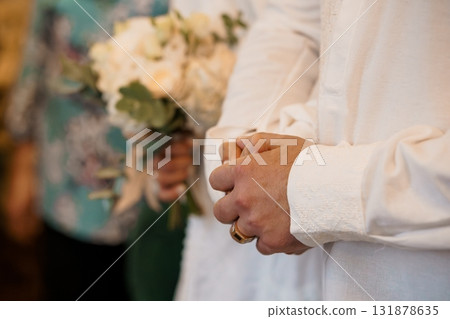 Wedding Ceremony Hands Clenched Together with Bouquet. White Flowers, Groom's Attire, Formal Event, Love, Joy, Blurred Background, Warm Tone. Wedding Ceremony Hands Clenched Together with Bouquet. White Flowers, Groom's Attire, Formal Event, Love, Joy, Blurred Background, Warm Tone. 131878635