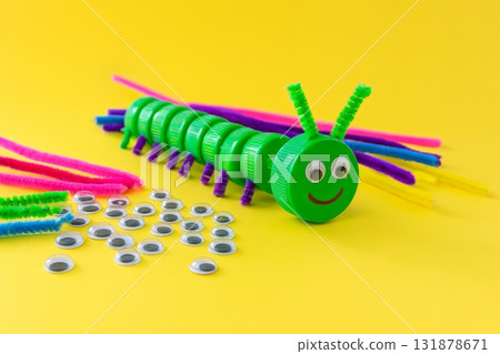 Caterpillar made from green plastic bottle caps with googly eyes and colorful chenille stems on yellow background. DIY craft project for kids. Flat lay composition for design and poster 131878671