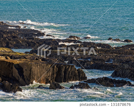 Waves crash against the jagged, dark rocks along the coast at Sheep Cove in Ireland. The sun shines on the water and the irregular stone formations. 131878676