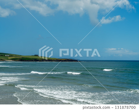 This is a view of the ocean on a sunny day in West Cork. Waves are moving toward the sandy beach. Grass and a house are visible along the coastline in Ireland. 131878700
