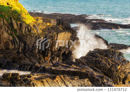 waves crash violently against the rugged, rocky coastline of South Ring in Ireland. The sun shines brightly on the rocks and water during the day. 131878712