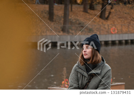 A young woman in a dark beanie and khaki jacket sits by a pond surrounded by autumn leaves and soft golden tones 131879431