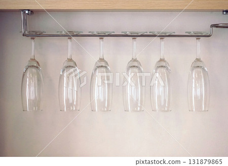 Skinny wine glasses hang on a stainless steel holder next to a wooden shelf on a white wall. 131879865