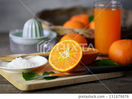 Half an orange is placed on a wooden chopping board. Placed on an old wooden table prepared for making orange juice, In the back there are oranges in the basket and the juicer container. Half an orange is placed on a wooden chopping board. Placed on an old wooden table prepared for making orange juice, In the back there are oranges in the basket and the juicer container. 131879876