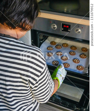 Vertical photo of woman taking hot baking tray filled with cookies from oven at home. Holiday baking scene, family kitchen moment, no face visible. Vertical photo of woman taking hot baking tray filled with cookies from oven at home. Holiday baking scene, family kitchen moment, no face visible. 131879912