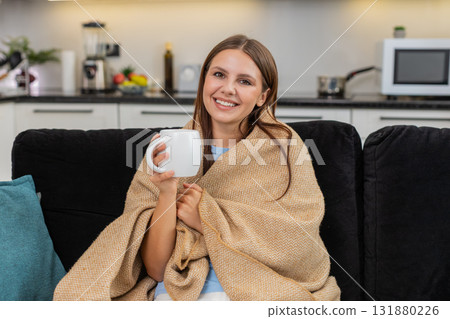 Young woman drinks herbal tea holds mug takes slow sip wraps in blanket relaxes and enjoys warm rest Young woman drinks herbal tea holds mug takes slow sip wraps in blanket relaxes and enjoys warm rest 131880226