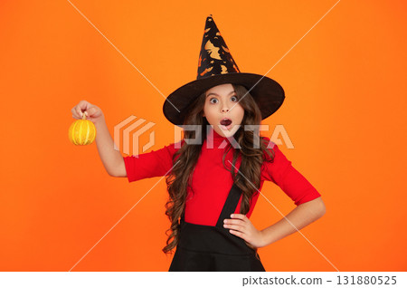 Halloween pumpkin in hand of surprised witch girl. Halloween witch girl isolated on orange. Pumpkin on Halloween. Witch All Hallows costume of girl. October 31 pumpkin. Costume party Halloween pumpkin in hand of surprised witch girl. Halloween witch girl isolated on orange. Pumpkin on Halloween. Witch All Hallows costume of girl. October 31 pumpkin. Costume party 131880525