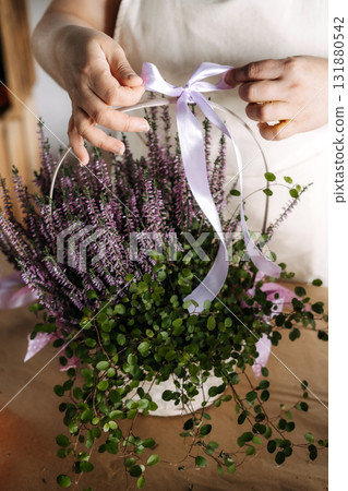 Person tying lavender ribbon around floral gift basket filled with heather and trailing greenery on a rustic table. Thanksgiving bouquet, handmade gift basket, seasonal flower gift, fall ribbon decor 131880542