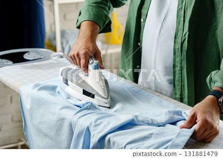 Man ironing a blue shirt in a well-lit room during the afternoon 131881479