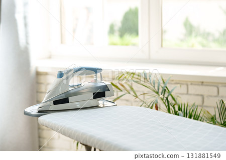 Iron rests on ironing board in bright laundry room with green plants and sunlight Iron rests on ironing board in bright laundry room with green plants and sunlight 131881549