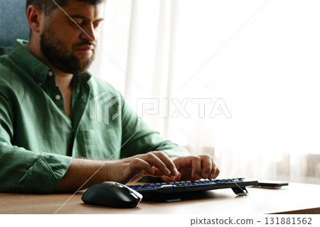 Man typing on keyboard while sitting at desk by window in a well-lit room 131881562