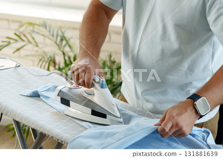 Person ironing a light blue shirt on an ironing board in a bright indoor space 131881639