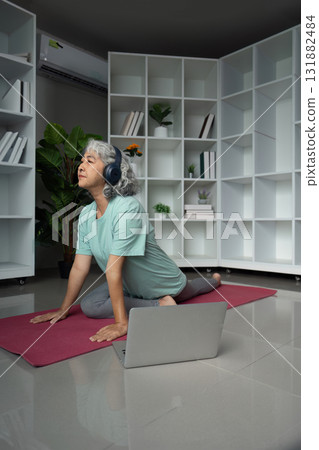 Senior woman practicing yoga with headphones and laptop in a serene indoor space 131882484