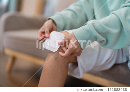 Young woman applying a bandage to her injured knee while sitting on a sofa in a soothing living room Young woman applying a bandage to her injured knee while sitting on a sofa in a soothing living room 131882488