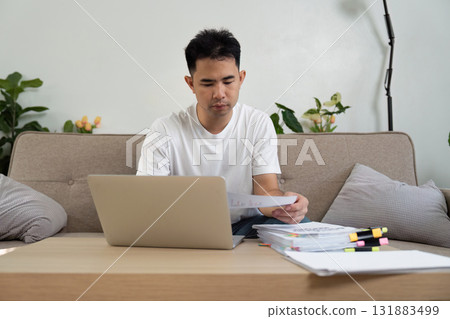 Young professional working on financial documents and laptop in a cozy home office. Young professional working on financial documents and laptop in a cozy home office. 131883499