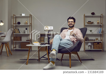 Happy man sitting in an armchair, using a projector, watching a movie, drinking tea and smiling Happy man sitting in an armchair, using a projector, watching a movie, drinking tea and smiling 131884189