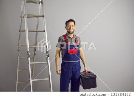 Portrait of a happy smiling repairman holding a tool box on a gray background near the ladder. 131884192