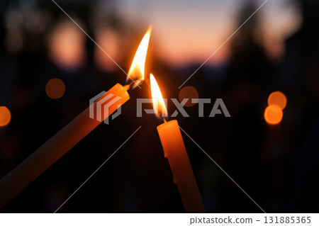 Two glowing candles being lit together outdoors at dusk, with a warm flame and blurred lights creating a peaceful evening atmosphere, AI Generative 131885365