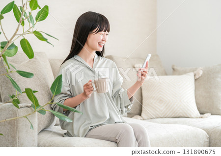 Young Asian woman sitting on the sofa at home drinking coffee and looking at her smartphone Young Asian woman sitting on the sofa at home drinking coffee and looking at her smartphone 131890675