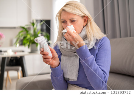 Middle aged blonde woman lying on couch at home, blowing her nose into a tissue and checking her temperature with a thermometer. Concept of flu, cold, fever, and home recovery. Middle aged blonde woman lying on couch at home, blowing her nose into a tissue and checking her temperature with a thermometer. Concept of flu, cold, fever, and home recovery. 131893732
