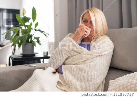 Middle aged woman wrapped in plaid sitting on couch and blowing her nose into a tissue. Concept of flu, covid, home rest, and self care during illness. Middle aged woman wrapped in plaid sitting on couch and blowing her nose into a tissue. Concept of flu, covid, home rest, and self care during illness. 131893737