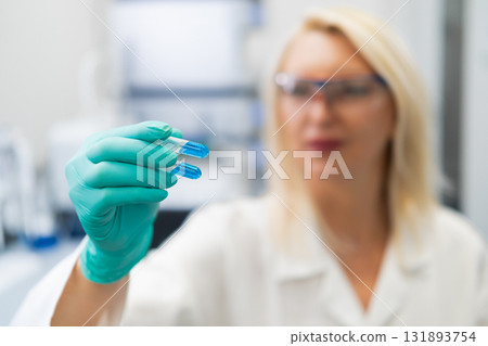 Blonde woman in lab coat studying blue liquid in test tube in chemistry lab. 131893754