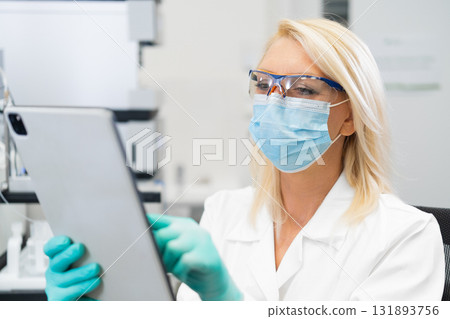 Middle aged female scientist wearing mask and lab coat using tablet in laboratory for research and analysis Middle aged female scientist wearing mask and lab coat using tablet in laboratory for research and analysis 131893756