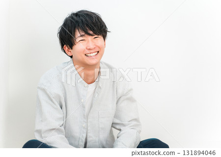 A young Asian man smiling while sitting in a living room with white walls A young Asian man smiling while sitting in a living room with white walls 131894046