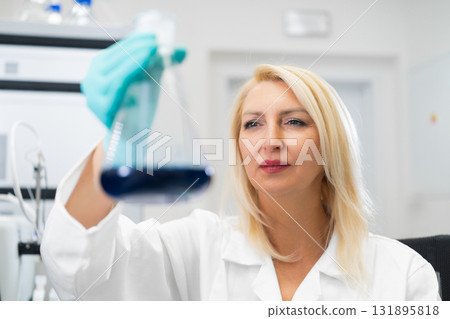 Blonde woman in rubber gloves examining a microbial culture in a flask while standing in a microbiology laboratory with HPLC and lab instruments in the background. Blonde woman in rubber gloves examining a microbial culture in a flask while standing in a microbiology laboratory with HPLC and lab instruments in the background. 131895818