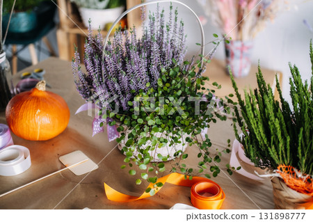Basket arrangement with purple heather and trailing greenery set beside a pumpkin and ribbons on a florist table. Local florist Thanksgiving visual, natural branding content 131898777