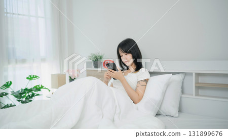 Young woman sitting in cozy bed, using smartphone and enjoying serene morning light in a minimalist bedroom atmosphere with plants and soft bedding Young woman sitting in cozy bed, using smartphone and enjoying serene morning light in a minimalist bedroom atmosphere with plants and soft bedding 131899676