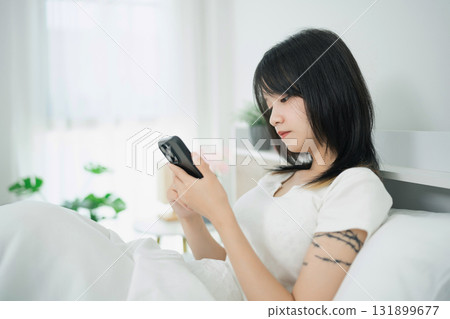 Young woman sitting in bed while using smartphone, wearing casual white clothing, surrounded by a bright and cozy bedroom environment with plants 131899677
