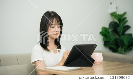 Young woman using digital tablet and stylus to create artwork or notes while seated at wooden table in cozy indoor environment with plant Young woman using digital tablet and stylus to create artwork or notes while seated at wooden table in cozy indoor environment with plant 131899683