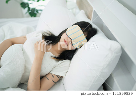 Relaxing woman sleeping peacefully with eye mask on white bedding in cozy bedroom with soft pillows and natural light streaming through window 131899829