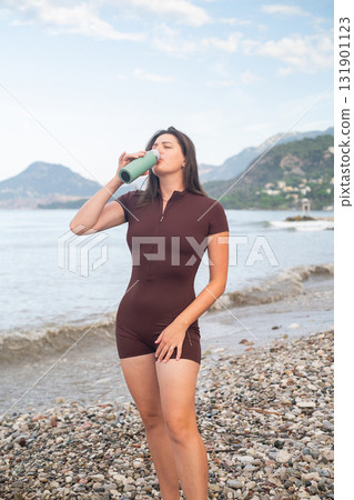 Woman restoring energy after workout by the sea fitness and health drinking water on the beach outdoor wellness Woman restoring energy after workout by the sea fitness and health drinking water on the beach outdoor wellness 131901123