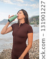 Woman in sportswear relaxing after morning workout by the sea drinking water from bottle outdoor fitness scene 131901227