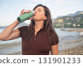 Active start of the day. Fit woman near the sea recovering after training and drinking water. 131901233