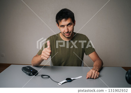 Friendly young man showing thumbs up gesture while charging smartwatch with portable power bank, expressing satisfaction with device and technology. Concept of healthy lifestyle. 131902932