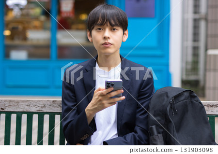 A male businessman in his twenties sitting on a bench and looking at his smartphone A male businessman in his twenties sitting on a bench and looking at his smartphone 131903098