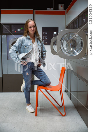 Woman standing with one leg on orange chair in self service laundromat with washing machines in background 131904539
