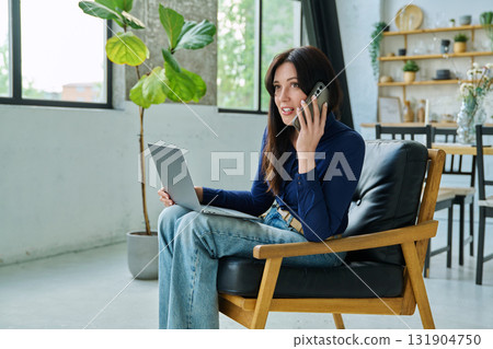 Positive young woman working at home with laptop talking on phone Positive young woman working at home with laptop talking on phone 131904750