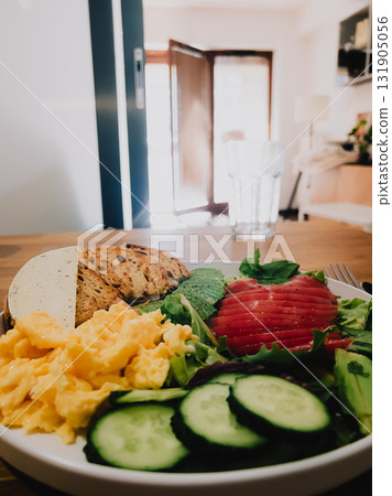 Scrambled eggs, sliced tomato, cucumber, avocado, lettuce, and toast arranged on white plate for nutritious breakfast, set on wooden table with glass of water. Balanced breakfast in homely atmosphere Scrambled eggs, sliced tomato, cucumber, avocado, lettuce, and toast arranged on white plate for nutritious breakfast, set on wooden table with glass of water. Balanced breakfast in homely atmosphere 131905056