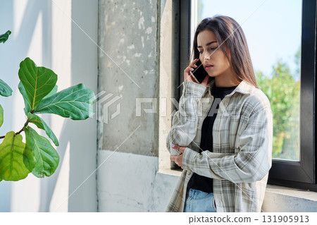 Serious teenage female standing at home talking on smartphone Serious teenage female standing at home talking on smartphone 131905913