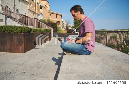 Casual Man Working on Laptop Outdoors Along Scenic Walkway With Railings Casual Man Working on Laptop Outdoors Along Scenic Walkway With Railings 131906566