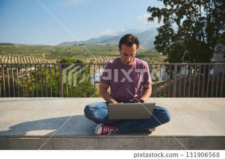 Casual Man Working on Laptop From Outdoor Terrace Above Vineyard Fields, Serene Mountain Views Casual Man Working on Laptop From Outdoor Terrace Above Vineyard Fields, Serene Mountain Views 131906568