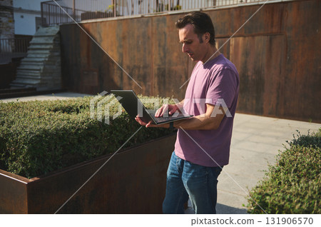 Focused Man Working on a Laptop Outdoors in Casual Pink T-Shirt and Jeans, Modern Urban Setting Focused Man Working on a Laptop Outdoors in Casual Pink T-Shirt and Jeans, Modern Urban Setting 131906570