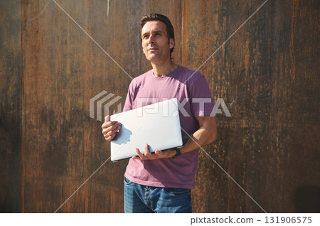 Professional Man With Laptop Standing Against A Rusty Wall, Casual Outdoor Work Vibe Professional Man With Laptop Standing Against A Rusty Wall, Casual Outdoor Work Vibe 131906575
