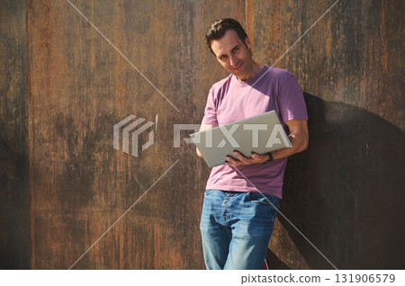 Casual Man With Laptop Leaning Against Rustic Wall Outdoors, Smiling, Everyday Tech Vibe 131906579